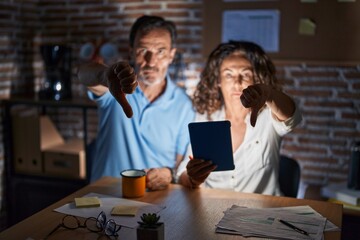 Middle age hispanic couple using touchpad sitting on the table at night looking unhappy and angry showing rejection and negative with thumbs down gesture. bad expression.