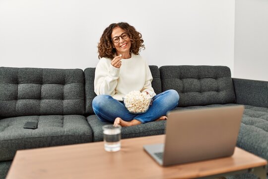 Mature Hispanic Woman Eating Popcorn Watching Tv Sitting On The Sofa At Home