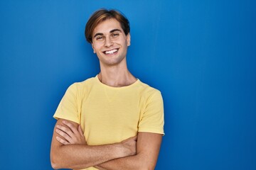 Young man standing over blue background happy face smiling with crossed arms looking at the camera. positive person.