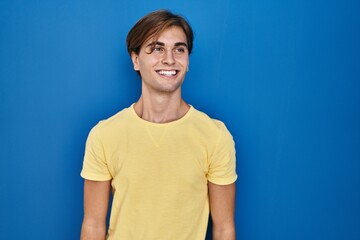 Young man standing over blue background looking away to side with smile on face, natural expression. laughing confident.