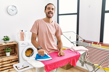 Young hispanic man ironing clothes at home sticking tongue out happy with funny expression. emotion concept.