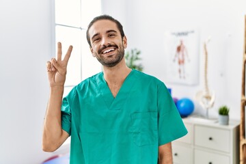 Young physiotherapist working at pain recovery clinic smiling with happy face winking at the camera doing victory sign. number two.