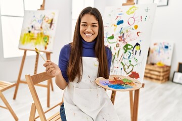 Young latin woman smiling confident holding paintbrush and palette at art studio