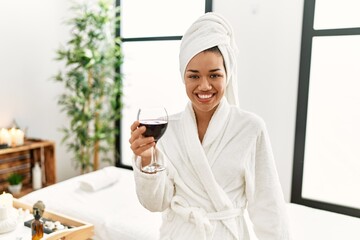 Young latin woman wearing bathrobe toasting with wine at beauty center