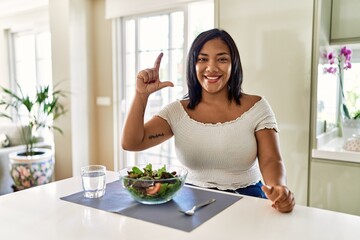 Obraz na płótnie Canvas Young hispanic woman eating healthy salad at home smiling and confident gesturing with hand doing small size sign with fingers looking and the camera. measure concept.