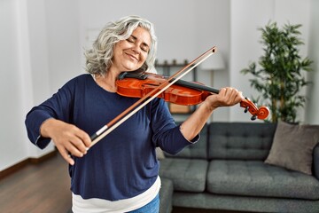 Middle age grey-haired artist woman playing violin standing at home.