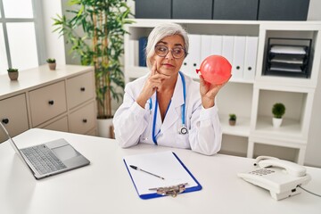 Middle age woman with grey hair wearing doctor uniform holding balloon serious face thinking about...