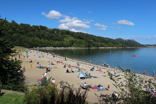 Sandy beach full of holiday makers, next to a calm body of water and surrounded by trees, with blue sky and white cumulus clouds overhead