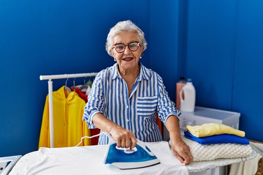 Senior grey-haired woman smiling confident ironing clothes at laundry room - Powered by Adobe
