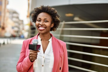Young african american woman journalist holding reporter microphone speaking and smiling to the...