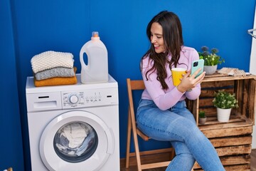 Young hispanic woman using smartphone and drinking coffee waiting for washing machine at laundry room