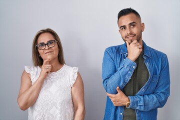 Hispanic mother and son standing together with hand on chin thinking about question, pensive expression. smiling and thoughtful face. doubt concept.