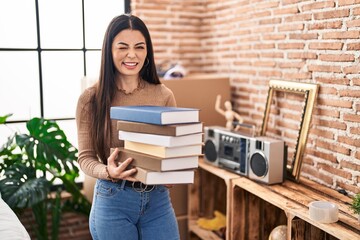 Young woman holding books at home winking looking at the camera with sexy expression, cheerful and...