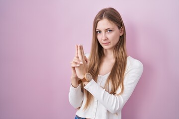 Young caucasian woman standing over pink background holding symbolic gun with hand gesture, playing killing shooting weapons, angry face