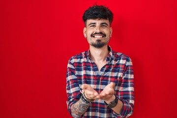 Young hispanic man with beard standing over red background smiling with hands palms together receiving or giving gesture. hold and protection