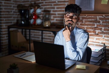 Young hispanic man with beard working at the office at night with hand on chin thinking about question, pensive expression. smiling with thoughtful face. doubt concept.