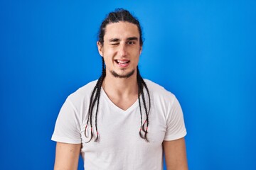 Hispanic man with long hair standing over blue background winking looking at the camera with sexy...