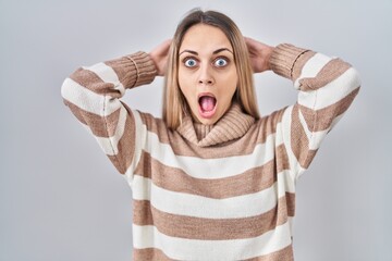Young blonde woman wearing turtleneck sweater over isolated background crazy and scared with hands...