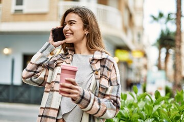 Young blonde woman holding coffee talking on the smartphone at street