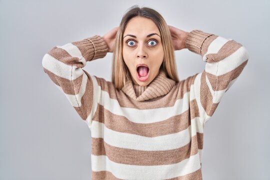 Young blonde woman wearing turtleneck sweater over isolated background crazy and scared with hands on head, afraid and surprised of shock with open mouth