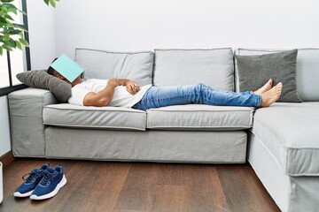 Young hispanic man lying on sofa sleeping with book on face at home