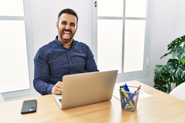 Young hispanic man with beard working at the office with laptop sticking tongue out happy with funny expression. emotion concept.
