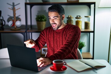 Young hispanic man with beard using computer laptop at night at home pointing with finger surprised ahead, open mouth amazed expression, something on the front