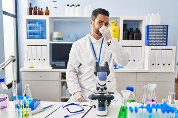 Young hispanic man with beard working at scientist laboratory smelling something stinky and disgusting, intolerable smell, holding breath with fingers on nose. bad smell