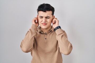Non binary person standing over isolated background covering ears with fingers with annoyed expression for the noise of loud music. deaf concept.
