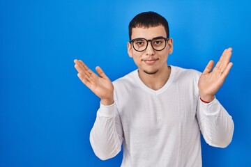 Young arab man wearing casual white shirt and glasses clueless and confused expression with arms and hands raised. doubt concept.