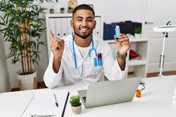 Young indian doctor holding medical asthma inhaler at the clinic smiling happy pointing with hand and finger to the side - obrazy, fototapety, plakaty