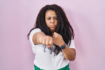 Plus size hispanic woman standing over pink background punching fist to fight, aggressive and angry attack, threat and violence