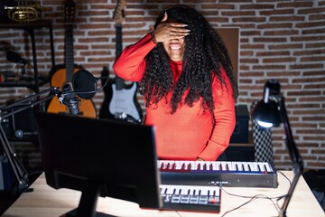 Plus size hispanic woman playing piano at music studio smiling and laughing with hand on face covering eyes for surprise. blind concept.