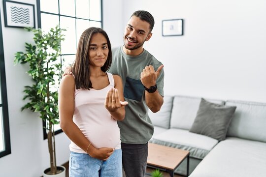 Young interracial couple expecting a baby, touching pregnant belly beckoning come here gesture with hand inviting welcoming happy and smiling