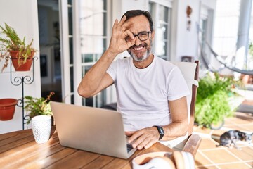 Middle age man using computer laptop at home doing ok gesture with hand smiling, eye looking through fingers with happy face.