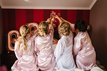 Young bridesmaids in silk robes drink champagne in hotel room. Beautiful females celebrating bachelorette party, sitting on bed. Back view.