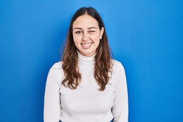 Obraz premium Young hispanic woman standing over blue background winking looking at the camera with sexy expression, cheerful and happy face.