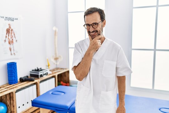 Middle age man with beard working at pain recovery clinic looking confident at the camera with smile with crossed arms and hand raised on chin. thinking positive.