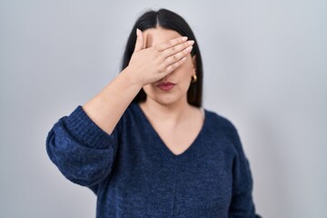 Young brunette woman standing over isolated background covering eyes with hand, looking serious and sad. sightless, hiding and rejection concept