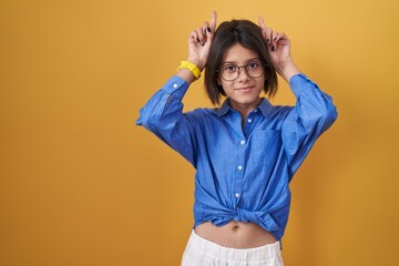 Young girl standing over yellow background doing funny gesture with finger over head as bull horns