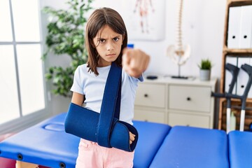 Young hispanic girl wearing arm on sling at rehabilitation clinic pointing with finger to the camera and to you, confident gesture looking serious