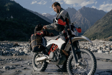 Male biker in long moto trip standing in mountains on mountain river near his motorcycle pulling something out of bags and luggage