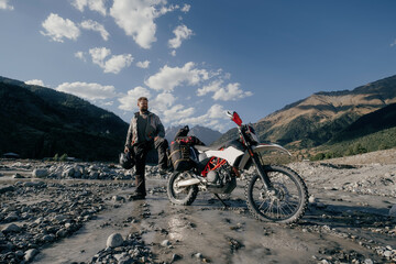 Naklejka premium Happy biker Man wearing beard and motorcycle clothes in long moto trip standing in mountains on river near his motorcycle