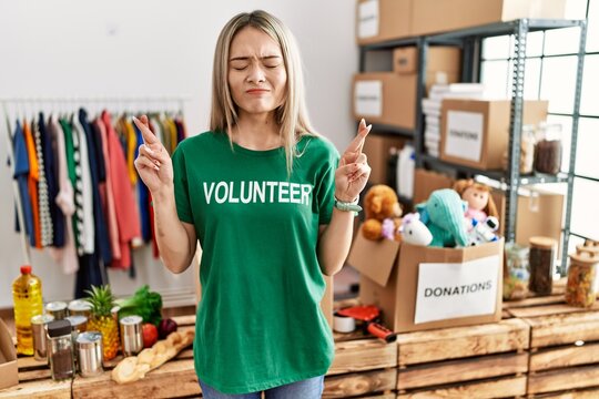 Asian young woman wearing volunteer t shirt at donations stand gesturing finger crossed smiling with hope and eyes closed. luck and superstitious concept.
