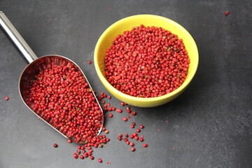 Pink pepper berries (Schinus terebinthifolia) in a yellow bowl and a stainless steel measuring spoon on a black surface, top view.