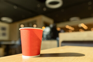 Cardboard red paper mug on a wooden table in café with blurred background lit by morning sun