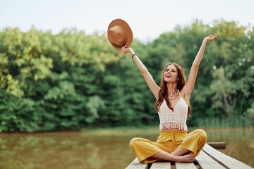 Hippie eco-activist woman traveler sits on a bridge by a lake with her arms outstretched with a hat and smiling sincerely