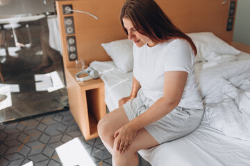 Fototapeta na wymiar Young woman sitting on bed and feeling knee pain, she massage her knee at home. Healthcare and medical concept. Pain in the knee.