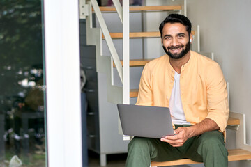 Smiling ethnic indian man wearing earphones sitting at home on stairs using laptop computer looking...