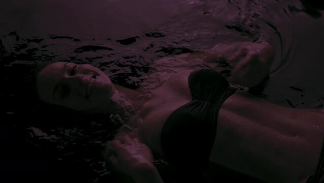 Beautiful fit sexy girl in bikini lying in dark water with colorful side light..